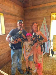 Two adults holding baby goats in the barn, with a young visitor standing between them