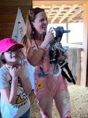 A laughing woman in pink overalls cradling a black baby goat beside a delighted child
