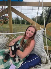 A woman on a hammock swing cuddling a black-and-white baby goat, pasture behind her