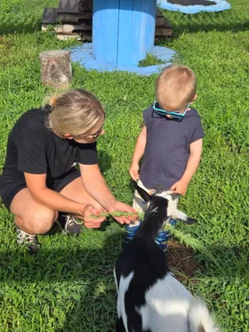 An adult kneeling beside a young child in sunglasses, offering a sprig of greenery to a goat