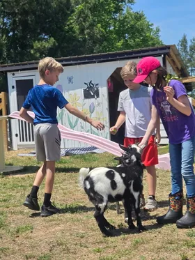 Three children feeding and petting two goats beside the flower-painted goat shed