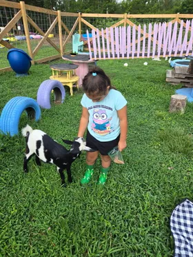 A child in green frog boots offering a handful of feed to a goat in the play yard