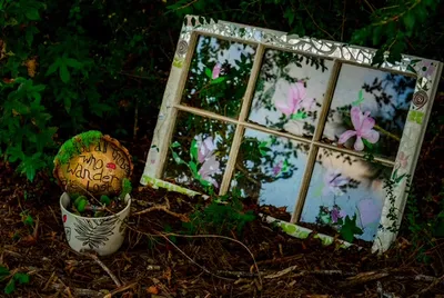 An old window frame painted with flowers, propped in the fairy garden beside a painted stone